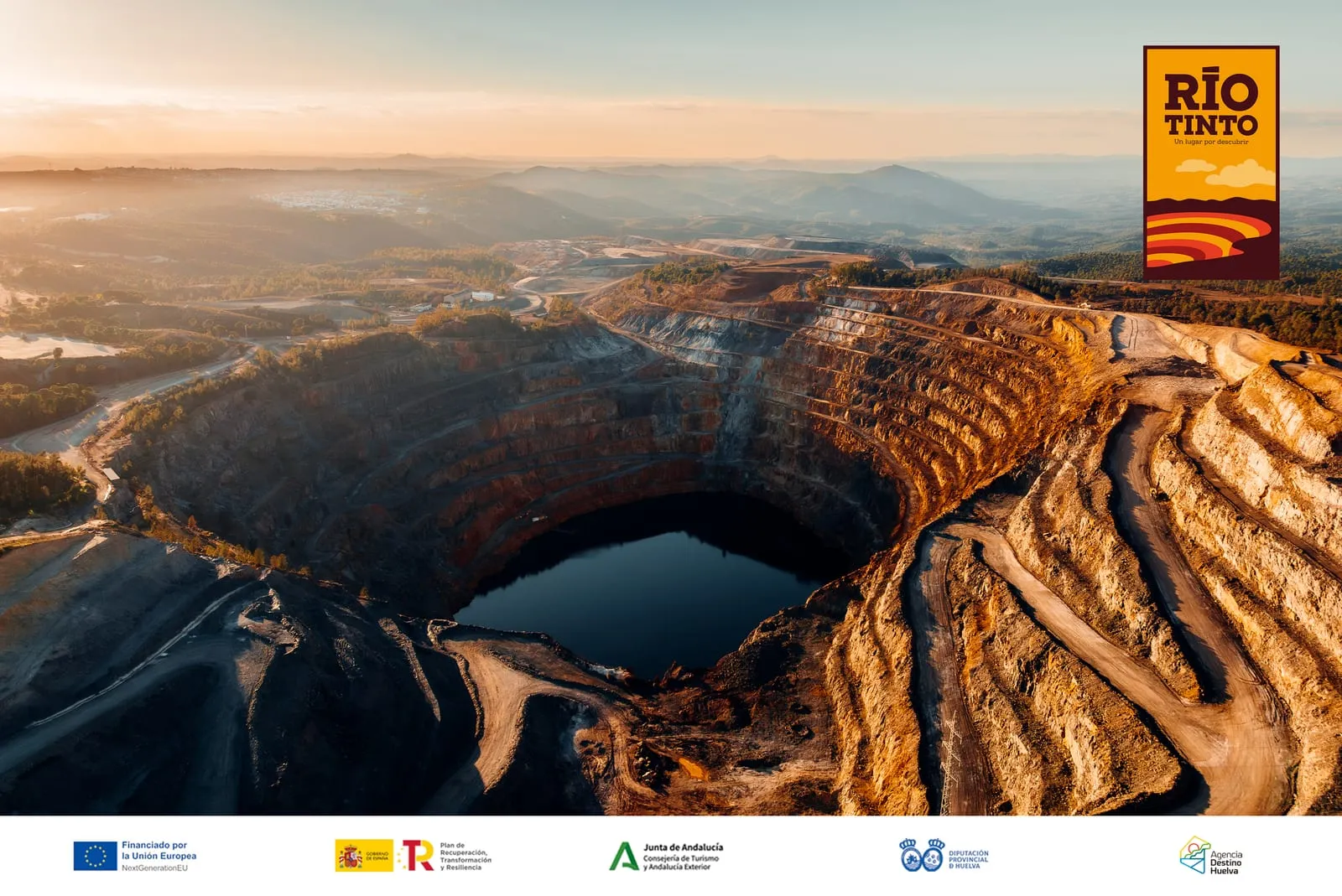 Vista aérea a contraluz de la corta minera de Riotinto al atardecer, con sus terrazas escalonadas y una laguna al fondo.