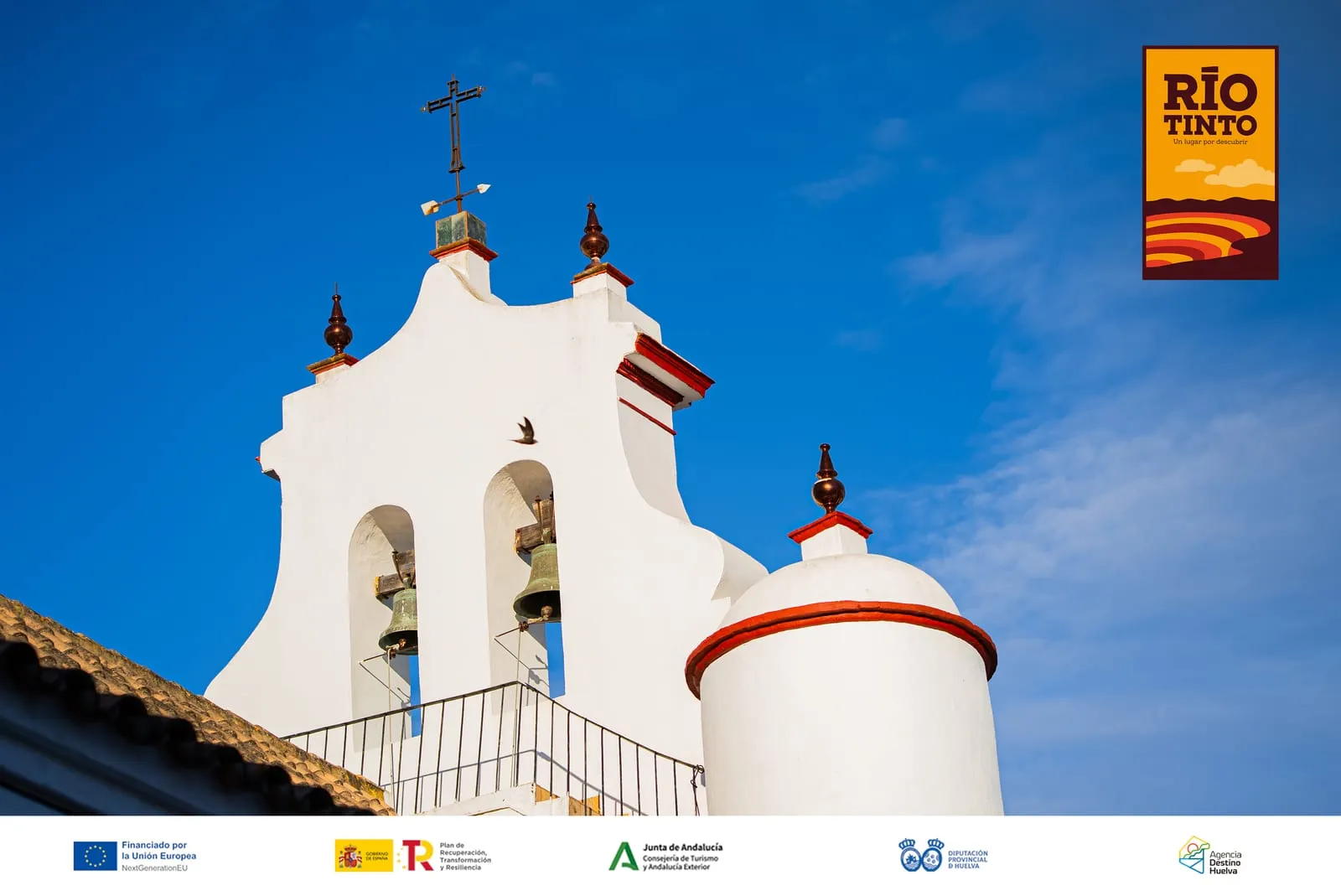 La espadaña blanca de la iglesia de Berrocal recortada contra un cielo azul, con sus campanas, la cruz y las veletas.