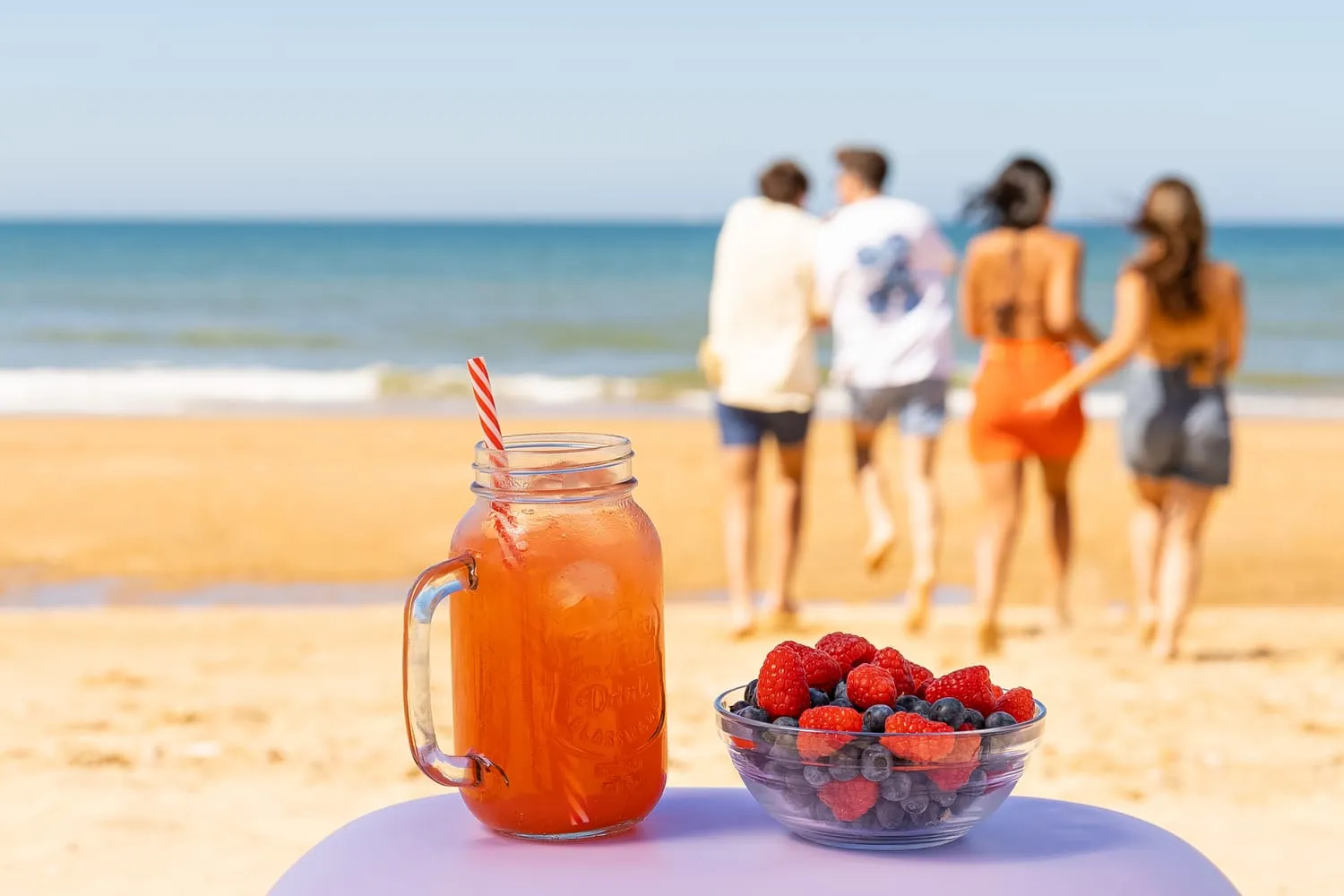 Bebida y bol de berries en primer plano, con un grupo de amigos caminando hacia el mar al fondo