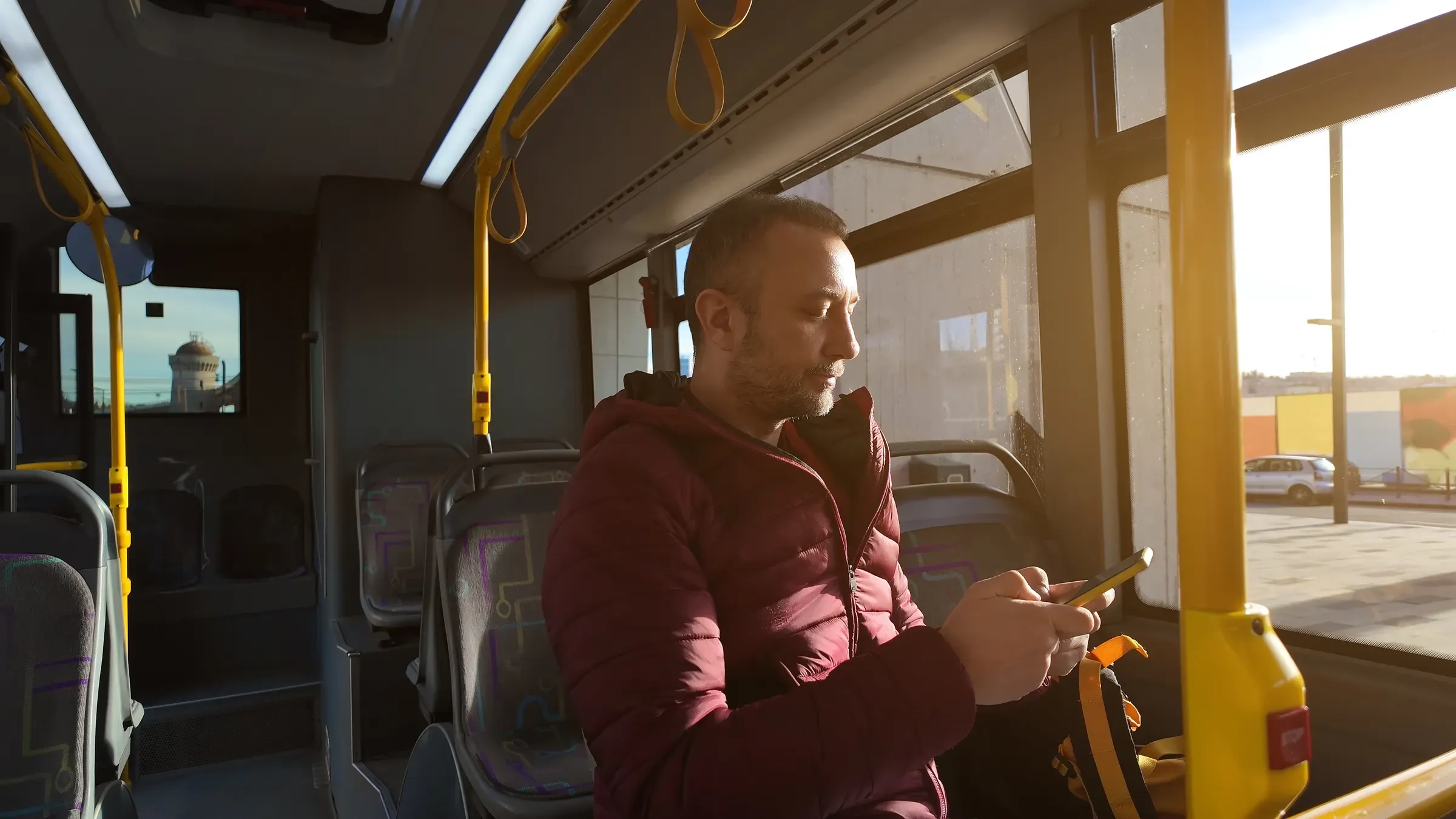Un hombre con plumífero granate mira el móvil sentado en un autobús urbano, con el sol de la tarde entrando por la ventanilla.