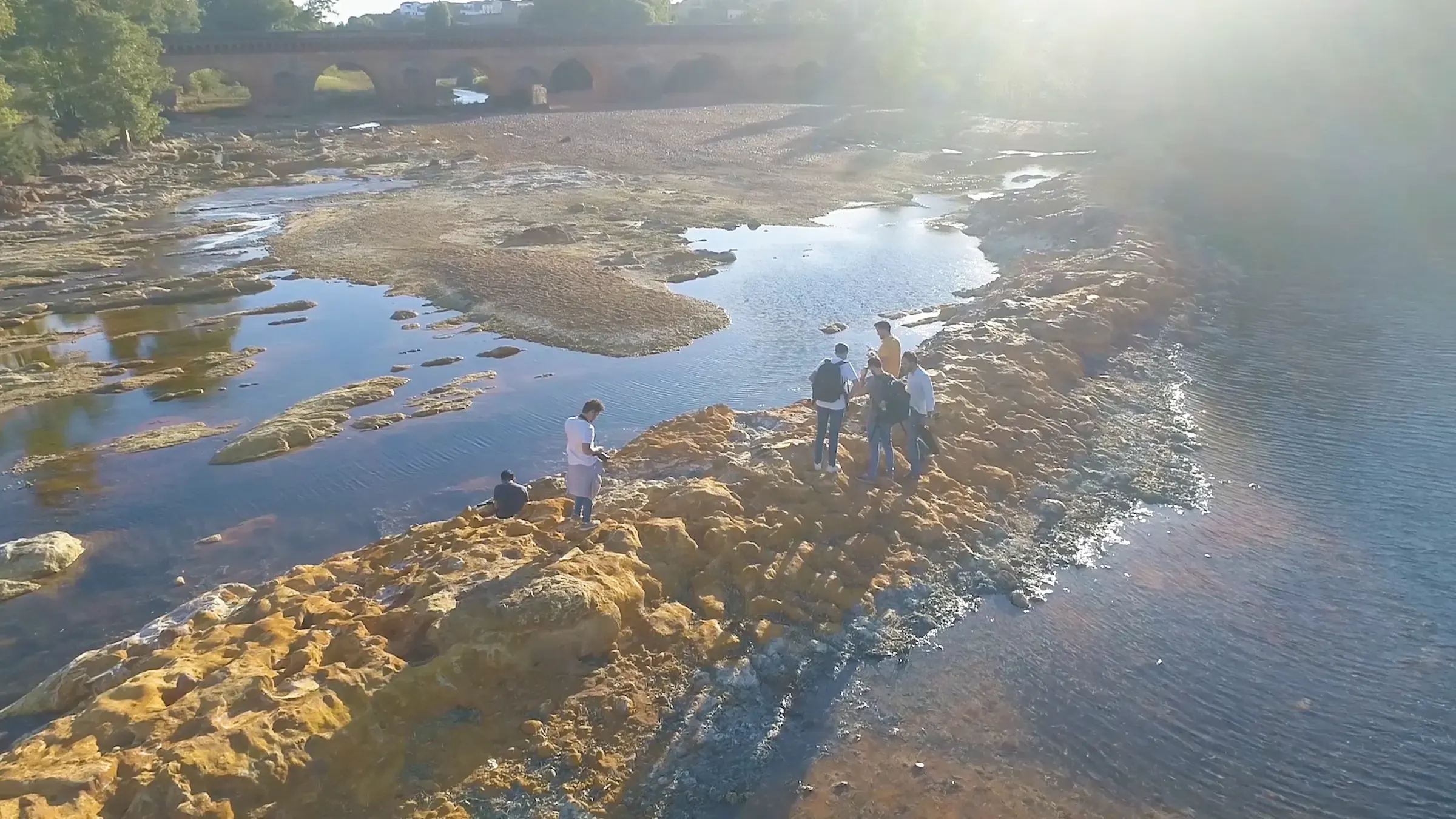 Vista aérea del equipo de Bis durante un rodaje, sobre las rocas de un río con un puente de piedra al fondo